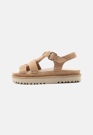 Beige ruskind sandal med justerbar ankelrem, guldspænde, tyk hvid rillet sål, og åben tå-design.