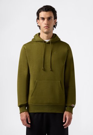 Giovane uomo che indossa un felpa verde oliva con tasca frontale e pantaloni neri, in piedi su uno sfondo leggero e semplice.