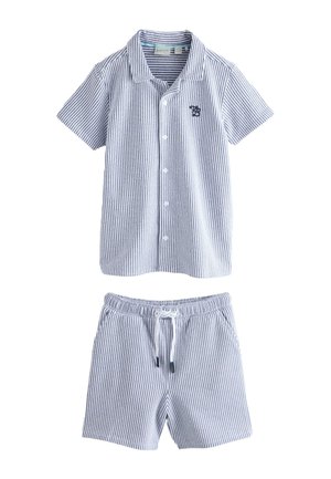 Lächelnder Junge geht, trägt kurzärmeliges gestreiftes Hemd und passende Shorts, weiße Socken und weiße Sneakers auf grauem Boden und Wandhintergrund.