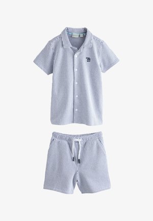 Lächelnder Junge geht, trägt kurzärmeliges gestreiftes Hemd und passende Shorts, weiße Socken und weiße Sneakers auf grauem Boden und Wandhintergrund.