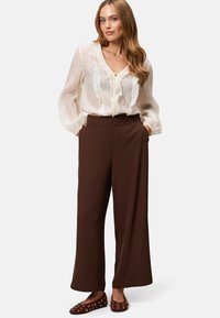 Flowy cream blouse with ruffled edges and tie detail, paired with high-waisted brown wide-leg trousers and brown slip-on shoes with studs.