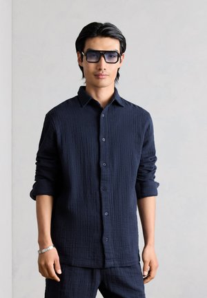 Mann mit schwarzen Haaren, der ein dunkelblaues, strukturiertes Hemd mit Knöpfen, passende Hose, Sonnenbrille und ein silbernes Armband trägt, steht vor einer hellgrauen Wand.