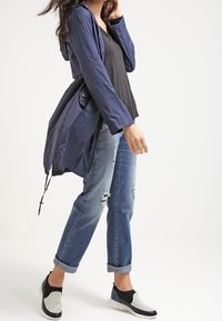 Veste légère bleu marine avec capuche, associée à un haut noir et un jean bleu déchiré. Chaussures à enfiler en maille noire et blanche.