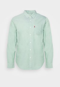 Chemise longue à manches longues vert clair avec col pointu et poche poitrine ornée d'une petite étiquette rouge sur le côté droit.