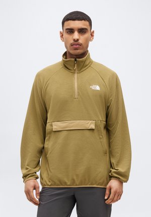 Mężczyzna w oliwkowo zielonej polarowej bluzie zapinanej na zamek do połowy szyi z przednią kieszenią i logo The North Face, stojący na jasnym, jednolitym tle.