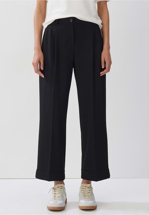 Personne portant un pantalon noir large court avec des ourlets retroussés, associé à des baskets blanches et un haut blanc, debout devant un fond uni.