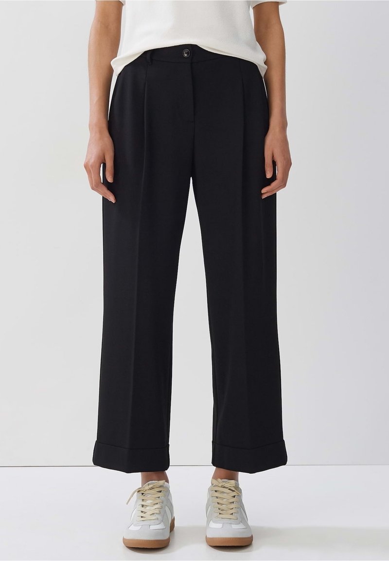 Personne portant un pantalon noir large court avec des ourlets retroussés, associé à des baskets blanches et un haut blanc, debout devant un fond uni.