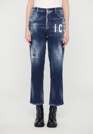 Jeans in denim blu scuro con finitura consumata, caratterizzati da grafiche bianche, dettagli strappati e un taglio dritto. Indossati con stivali neri.