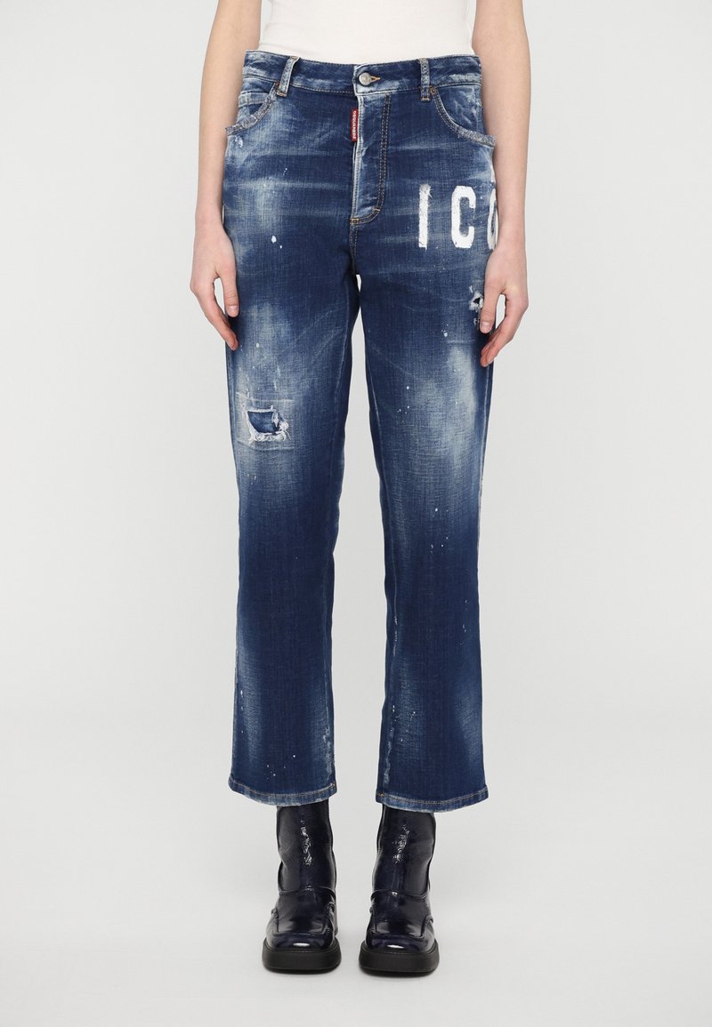 Jeans in denim blu scuro con finitura consumata, caratterizzati da grafiche bianche, dettagli strappati e un taglio dritto. Indossati con stivali neri.