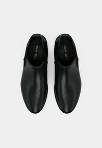 O pereche de pantofi din piele negri, tip slip-on, cu vârfuri rotunjite, pe un fond deschis, cu numele mărcii vizibil în interior.