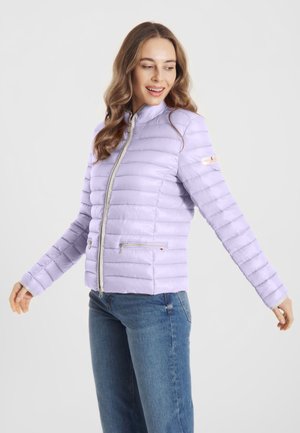 Kobieta w jasnofioletowej pikowanej kurtce z kieszeniami na zamek, stojąca z lekko wyciągniętymi ramionami, uśmiechająca się i patrząca w bok.