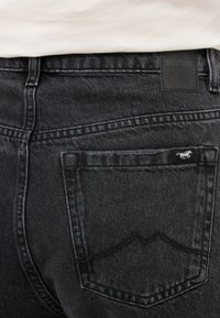 Černé džíny z denimu s mírně texturovaným povrchem. Na pase je štítek s nápisem "MUSTANG" a malé logo na zadní kapse.