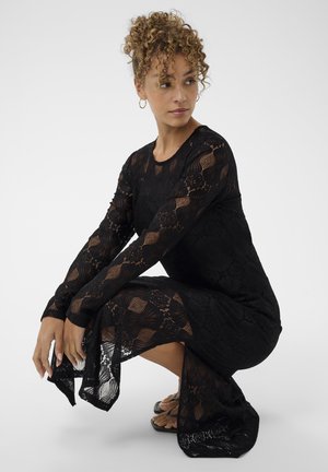 Vestido negro de encaje con mangas largas, que presenta patrones geométricos y un dobladillo acampanado. La modelo está en cuclillas, mostrando la textura transparente del vestido.