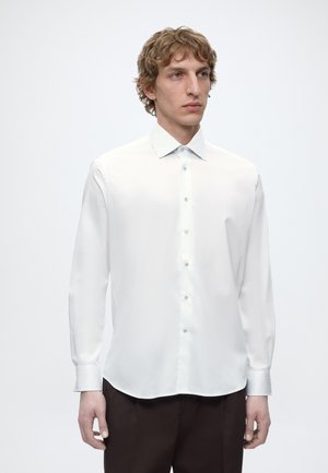 Mann mit lockigem blondem Haar, der ein weißes langärmeliges Hemd und dunkle Hosen trägt, steht vor einem schlichten weißen Hintergrund.