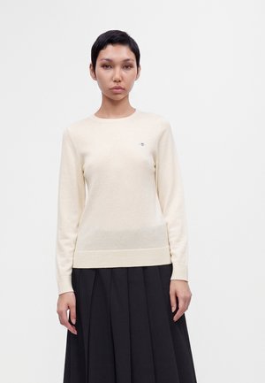 Femme aux cheveux courts et foncés portant un pull beige clair à manches longues et une jupe plissée noire, debout devant un fond blanc uni.