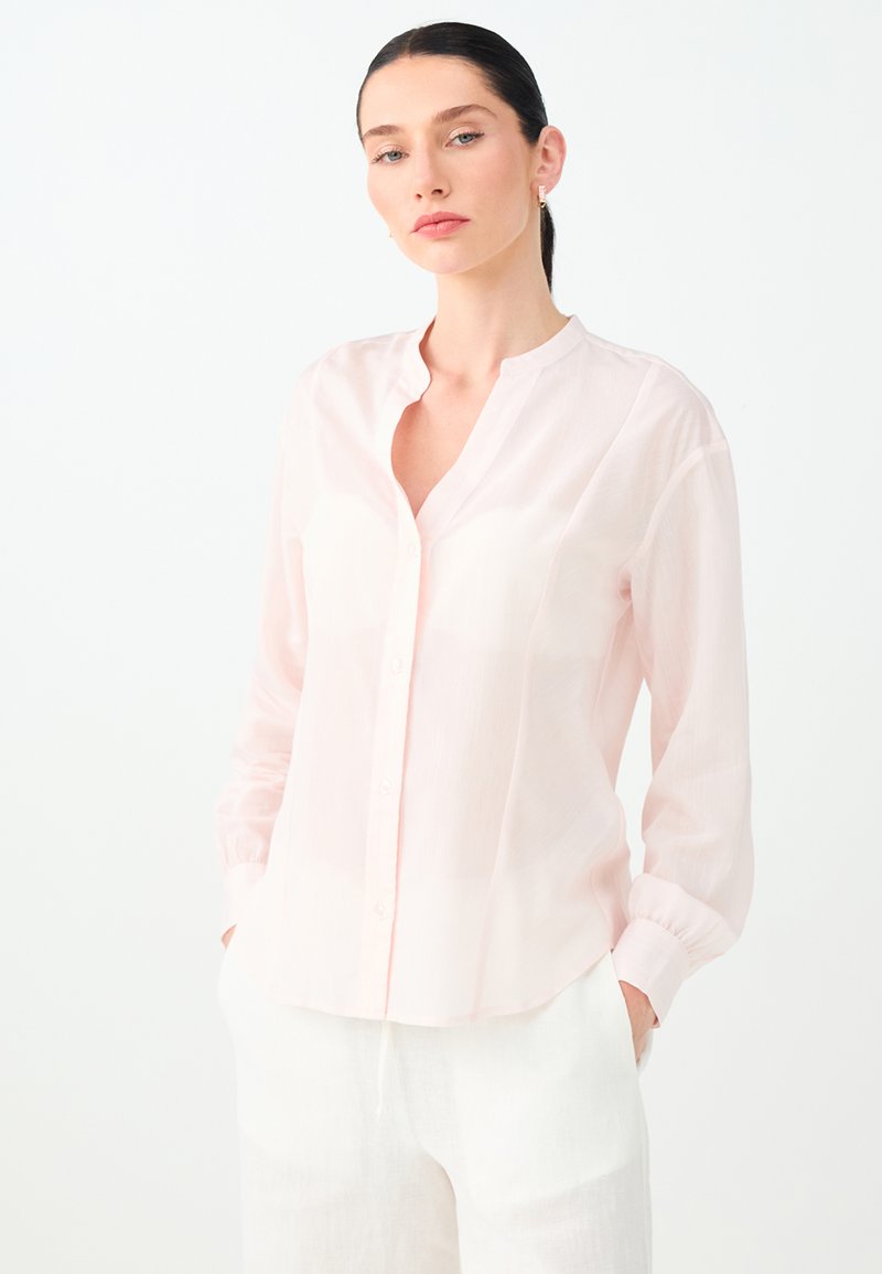 Femme aux cheveux foncés attachés, portant un chemisier rose clair boutonné et un pantalon blanc, debout les mains dans les poches devant un fond blanc.