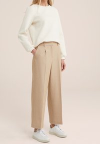 Nainen, joka on pukeutunut kermaiseen, teksturoituun pitkähihaiseen puseroon, beigeihin korkeavyötäröisiin leveälahkeisiin housuihin ja valkoisiin lenkkareihin, seisoo yksi käsi taskussa.