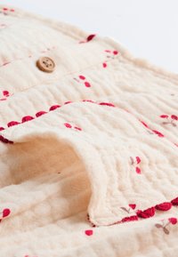 Tissu beige clair avec une surface texturée, ornée d'une broderie florale rouge et rose et d'une fermeture à bouton marron.