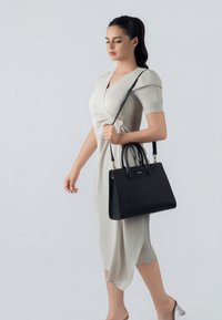 Borsa nera con texture, manici doppi e tracolla staccabile, tenuta da una modella con un vestito grigio chiaro, su uno sfondo neutro.