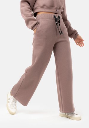 Donna che indossa pantaloni sportivi larghi a vita alta color malva con coulisse nere, abbinati a sneakers bianche, in piedi con una gamba avanti.