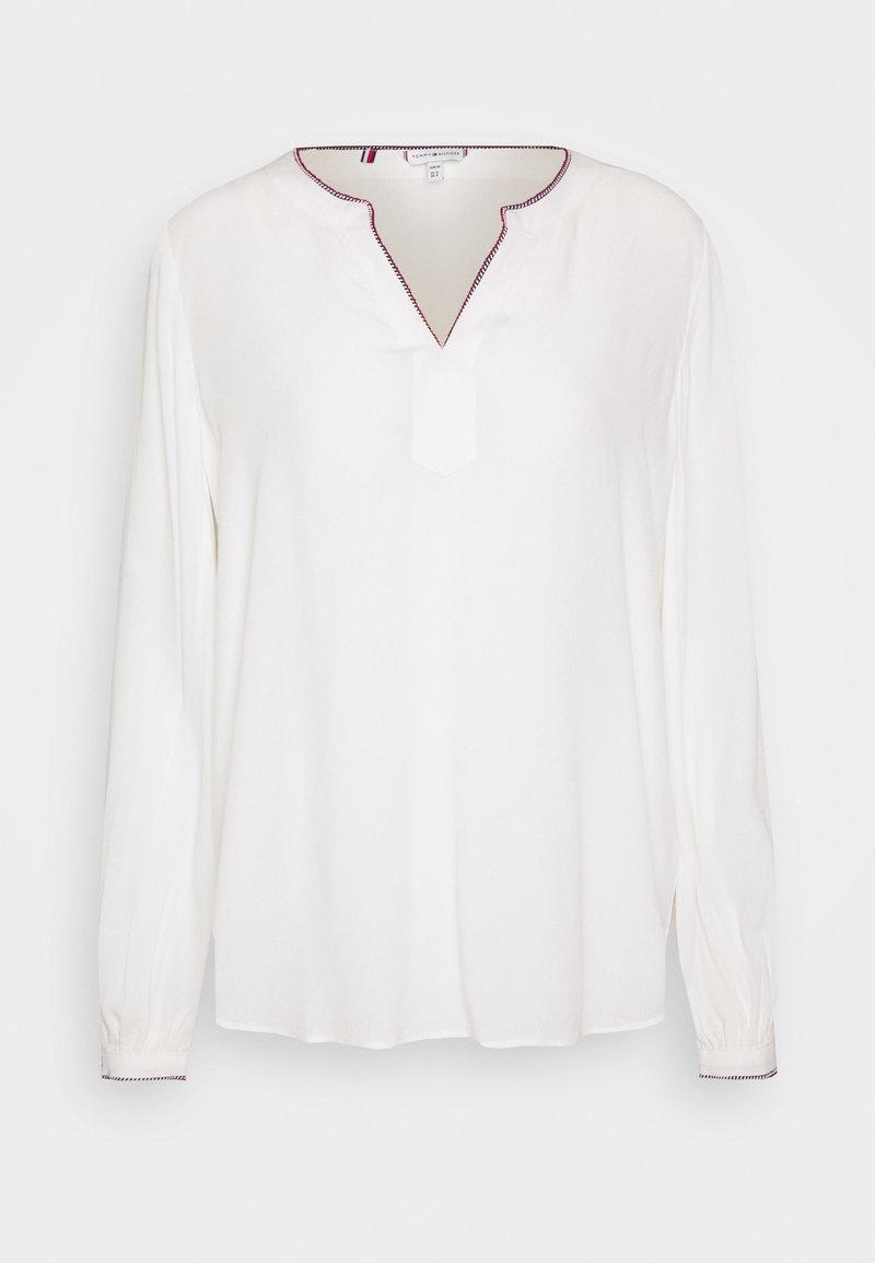 Blusa bianca a maniche lunghe con scollo a V e un piccolo taglio, decorata da cuciture rosse lungo il colletto e i polsini. Tessuto morbido.