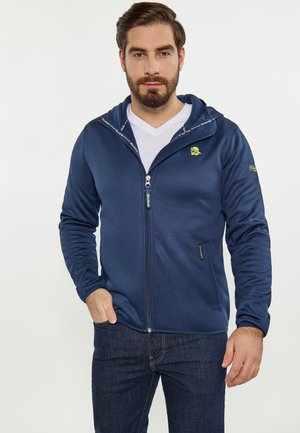 Uomo che indossa una felpa con cappuccio blu navy con zip sopra una t-shirt bianca e jeans scuri, in piedi con una mano in tasca contro uno sfondo semplice.