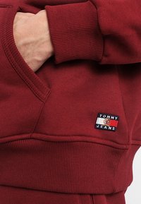 Felpa maroon con polsini e orlo a coste, che presenta un piccolo logo ricamato "Tommy Jeans" in rosso, bianco e blu sulla tasca frontale.