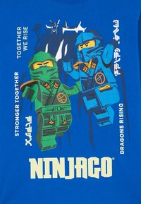 Modrá tričko s dvoma stylizovanými ninja postavami v zelenej a modrej farbe. Text znie "NINJAGO," "SPOLU SOME SILNÍ" a "DRAGONY RASTÚ."