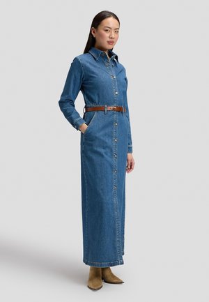 Femme debout portant une longue robe en jean bleue boutonnée, une ceinture marron et des bottines couleur crème sur un fond gris uni.