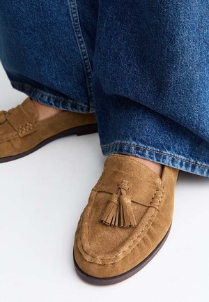 Mocasines de ante marrón con borlas, combinados con vaqueros azules, mostrados sobre fondo blanco.