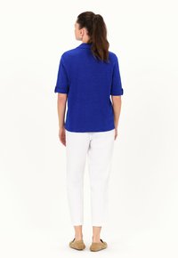 Polo shirt blu con texture e maniche arrotolate, abbinato a pantaloni bianchi a sigaretta e scarpe slip-on marroni, vista da dietro.