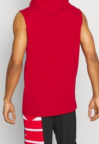 Punainen hihaton huppari, jolla on sileä pinta ja takaosassa huppu sekä suora helma, yhdistetty mustiin shortseihin.