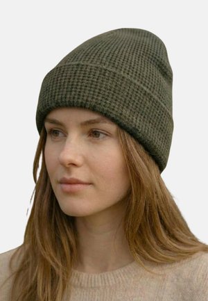 Junge Frau mit langen braunen Haaren, die eine strukturierte olivgrüne Strickmütze und einen beigen Pullover trägt, schaut leicht nach links.