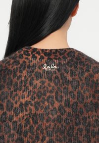 Mulher com cabelo preto longo, vestindo um suéter felpudo de padrão onça marrom e preto, com "lala Berlin" bordado nas costas próximo ao decote.