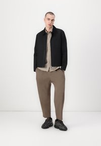 Modelo con una chaqueta negra con cremallera, camiseta beige a rayas y pantalones anchos marrones, combinados con zapatillas texturizadas negras. Fondo blanco liso.