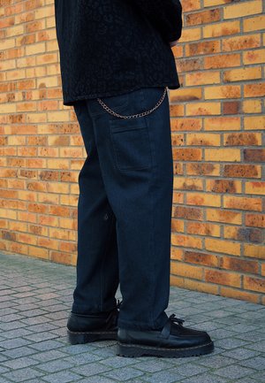 Osoba nosząca czarne skórzane buty, ciemnoniebieskie spodnie z boczną kieszenią i łańcuszkiem, stojąca na wyłożonym płytkami chodniku przy żółtej ceglanej ścianie.