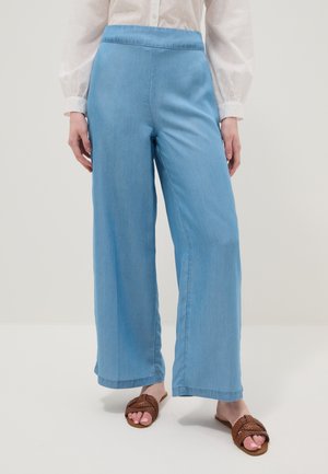 Mujer con pantalones anchos de color azul claro, blusa blanca transparente y sandalias marrones tejidas, de pie sobre un fondo neutro.
