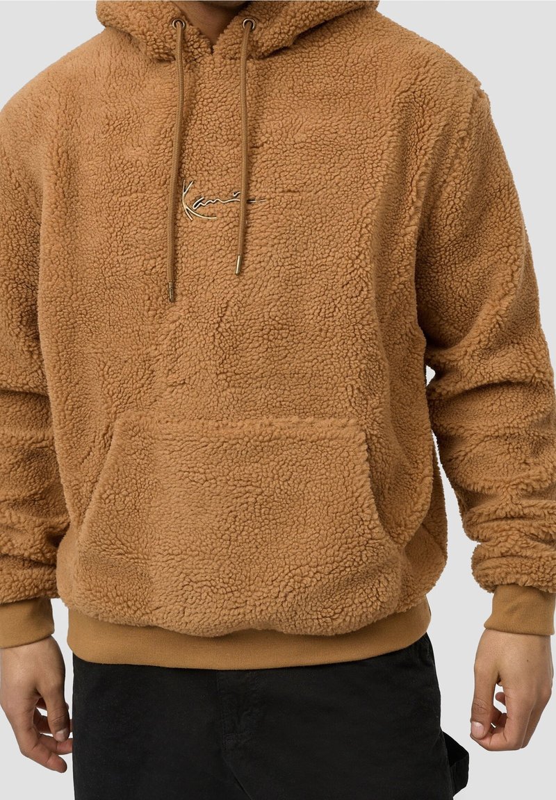 Sweat à capuche en polaire sherpa marron avec poche avant et cordons, porté par une personne avec un pantalon noir, montré des épaules aux hanches.