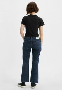 T-shirt noir ajusté, jean en denim bleu foncé taille haute avec poches arrière plates, légère décoloration en rayures, et baskets noires hautes. Design simple.