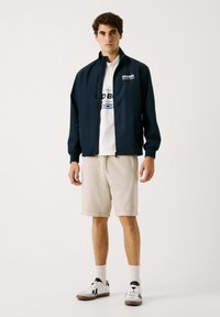 Veste coupe-vent marine avec des accents bleus, polo blanc avec logo, shorts beiges et baskets blanches avec des détails noirs et marron.
