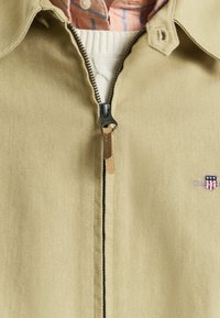 Beige jakke med lynlås foran, krave og tekstureret stof. Har en læder lynlås træk og et lille logo patch på brystet.