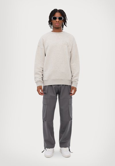 Lichtgrijze sweatshirt, ontspannen pasvorm met ribgebreide manchetten. Donkergrijze cargobroek met meerdere zakken. Witte sneakers en zwarte zonnebril.