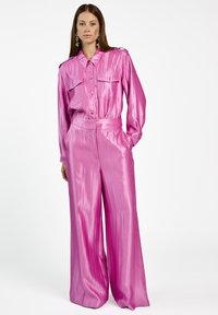 Glanzende roze blouse met knoopsluiting aan de voorkant en twee borstzakken, gecombineerd met wijde roze broek in een strakke satijnen stof.