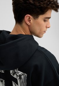 Sweat à capuche noir avec un grand motif métallique au dos. Doté d'une capuche avec cordon de serrage et de poignets côtelés. Tissu doux et texturé.