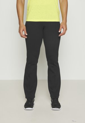 Personne portant un pantalon noir ajusté, des baskets noires à semelles blanches et un t-shirt jaune, debout sur un sol et un mur clairs unis.