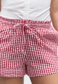 Κοντινό πλάνο κόκκινων και λευκών καρό σορτς με κορδόνι δεμένο σε φιόγκο, φορεμένα με λευκό πουκάμισο μέσα από το παντελόνι.