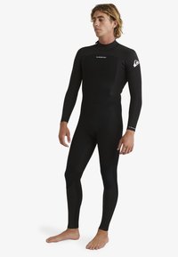 Full-body wetsuit in black neoprene, featuring long sleeves, high neck, and seamless panels for flexibility. Logo accents on shoulder.