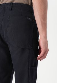 Zwarte cargobroek met meerdere zakken, een platte tailleband en een gestructureerde afwerking. Zichtbare stiksels en een klein patch-detail op de achterzak.