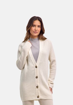 Femme portant un long cardigan en maille crème avec quatre boutons par-dessus un col roulé gris clair, debout sur un fond blanc uni.
