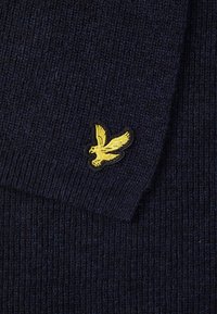 Vêtement en tricot de couleur bleu marine avec une texture côtelée et un col à rabat. Présente un logo d'aigle brodé jaune sur fond noir.